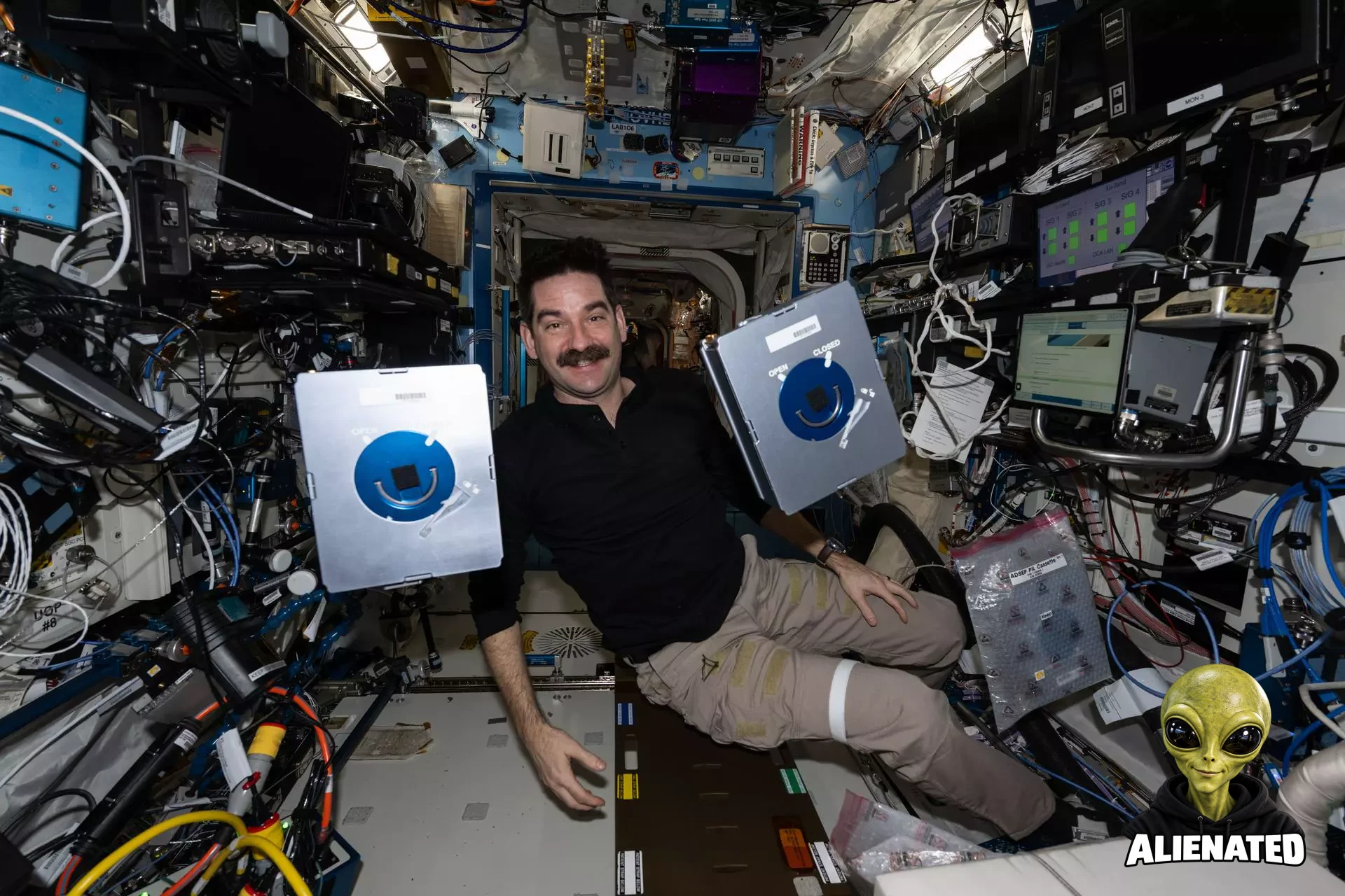 NASA astronaut Jack Hathaway floats and smiles at the camera, his face framed by two metallic silver boxes. Around him, the International Space Station’s walls are densely packed with wires, tubes, computer screens, and other hardware.