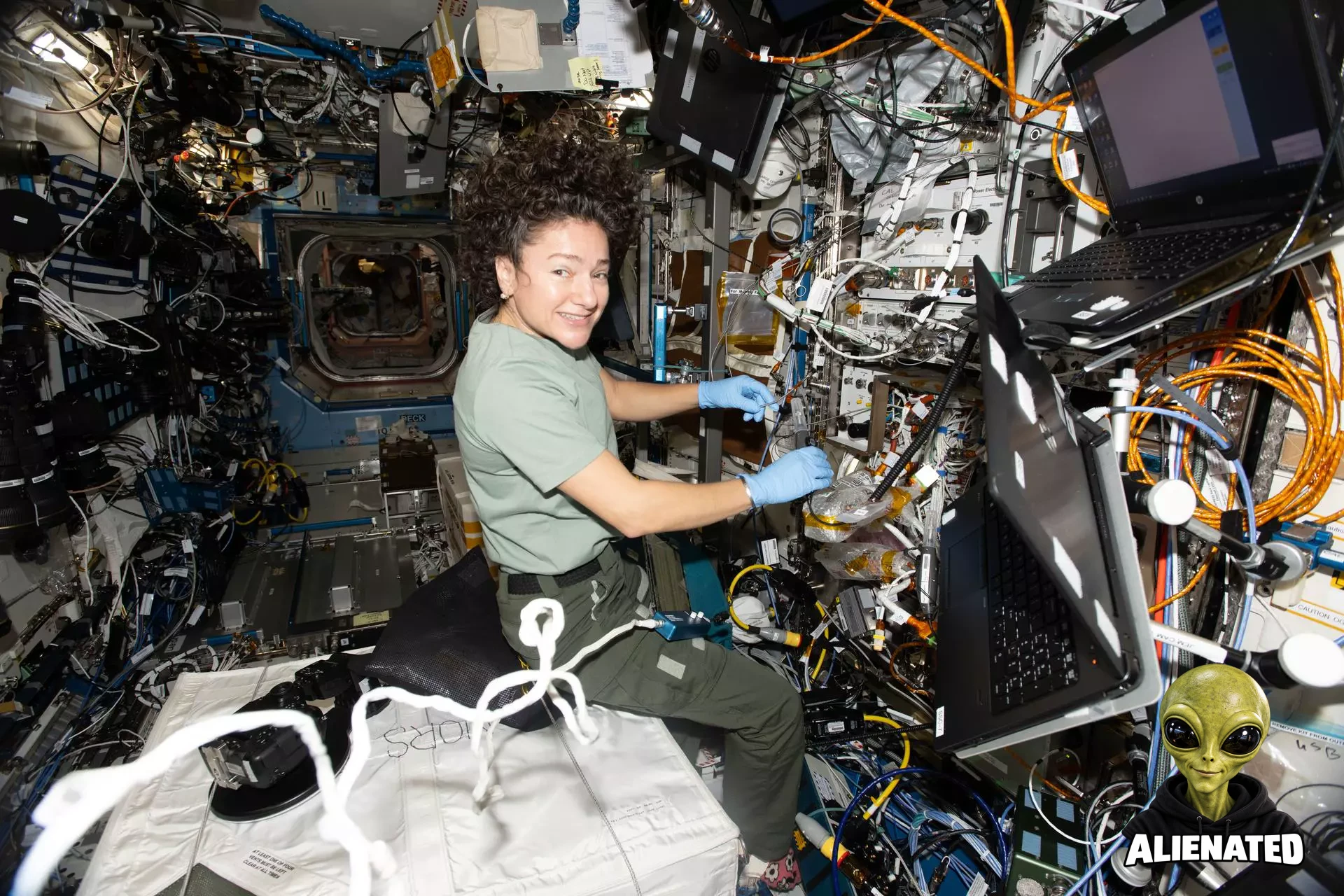 NASA astronaut Jessica Meir wears blue gloves and smiles at the camera while working with equipment aboard the International Space Station. Cables, tubes, and hardware cover the wall around her, and two laptops are mounted to her right. She rests on a large white padded container, giving her a seated appearance in microgravity.