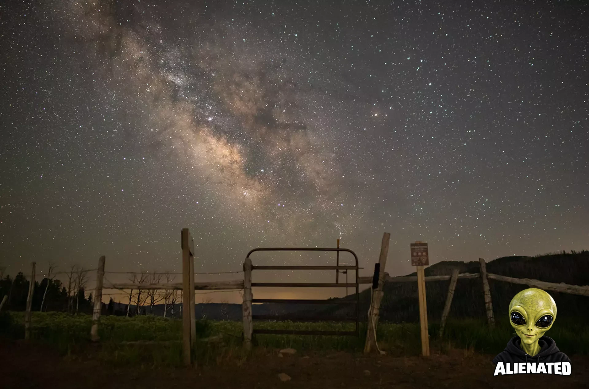 The bright, dusty band of the Milky Way galaxy stretches across a star-filled night sky. In the foreground, a rustic wooden fence and a closed metal gate stand on a grassy field, with dark hills silhouetted in the distance.
