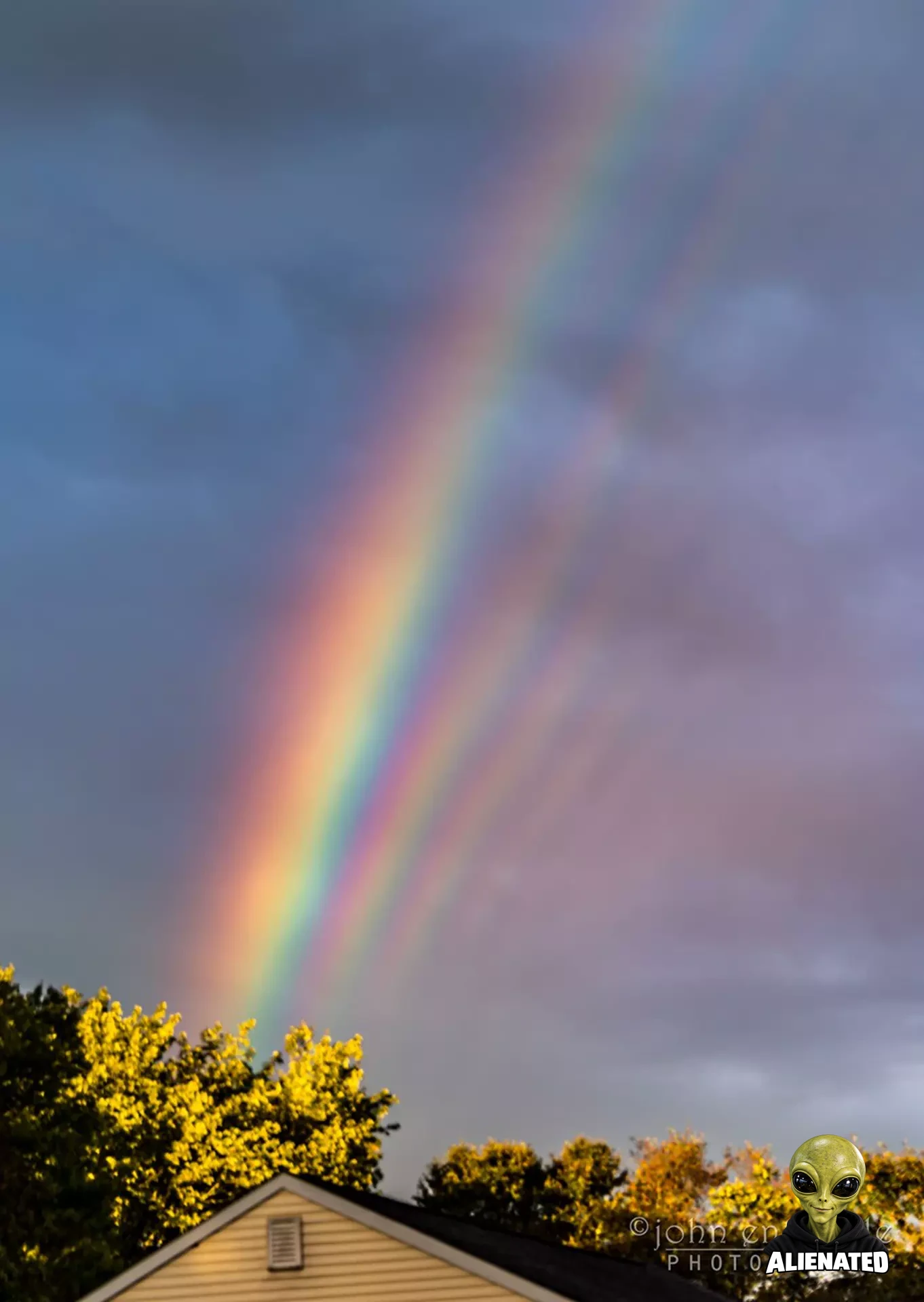 The top of a house is shown beneath a dark and cloudy sky. Many rainbow stripes are seen above the house.