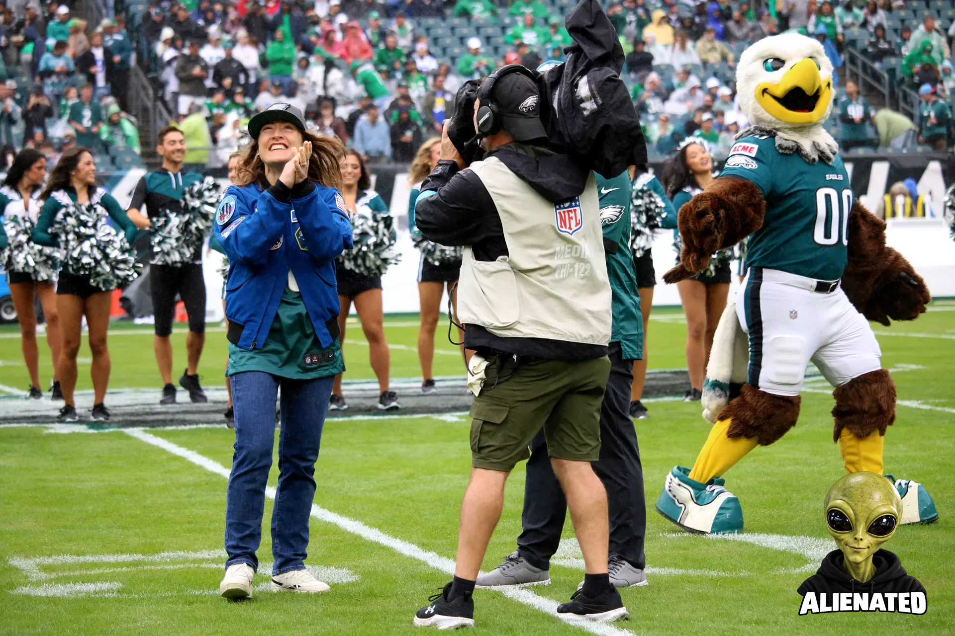 NASA astronaut Christina Koch (left, facing the camera) claps on a football field. An NFL cameraperson films her. The Philadelphia Eagles mascot is on the field as well (right), along with a line of cheerleaders in the background. Fans can be seen in the stands.