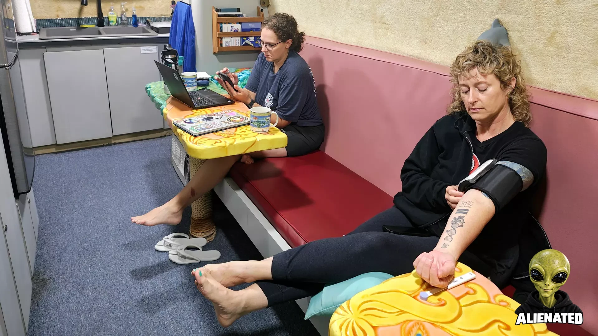 two women sit on a red bench in a small kitchen with blue carpet